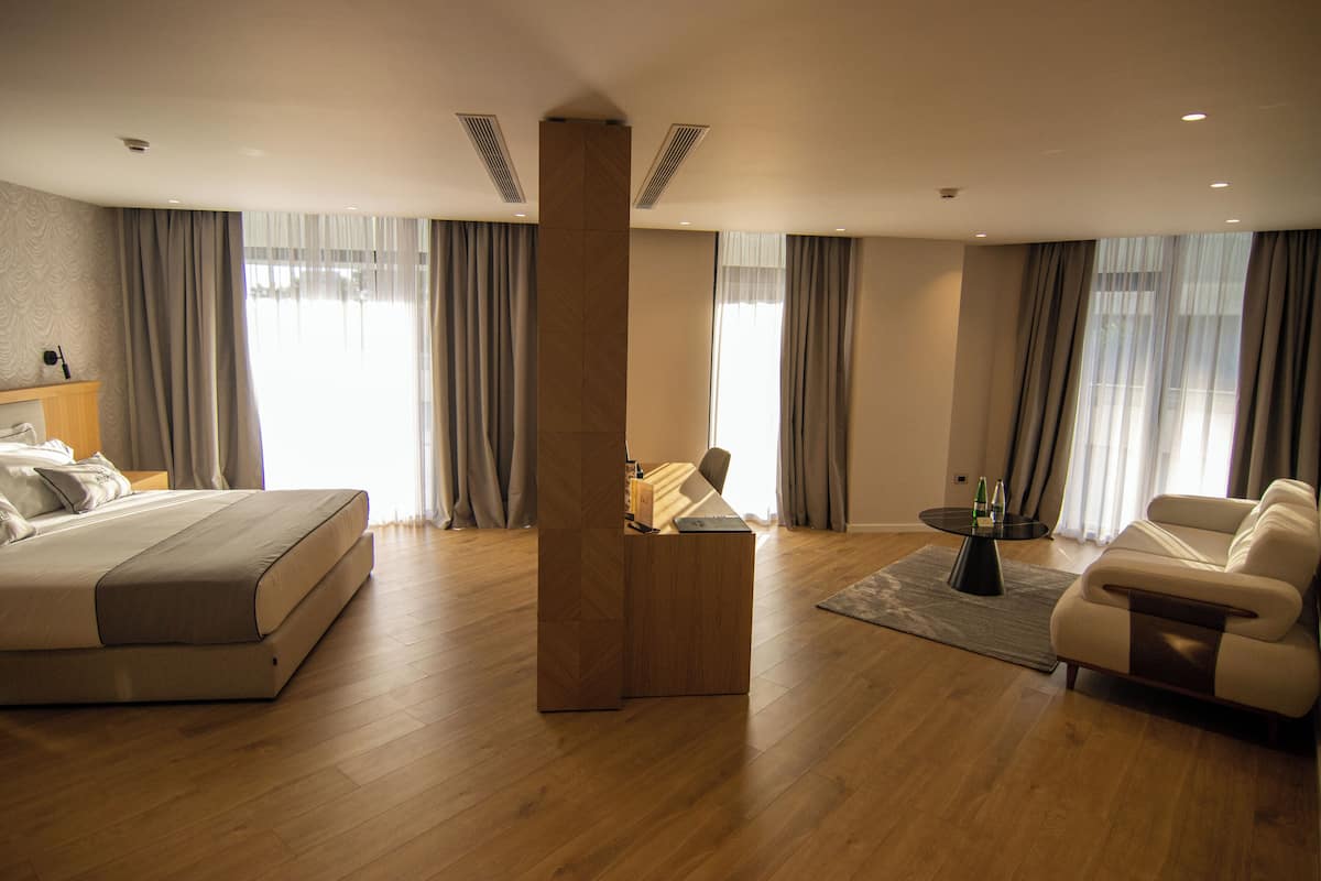 A modern hotel room with a large bed, a desk, a sofa, and a view of the city through large windows.