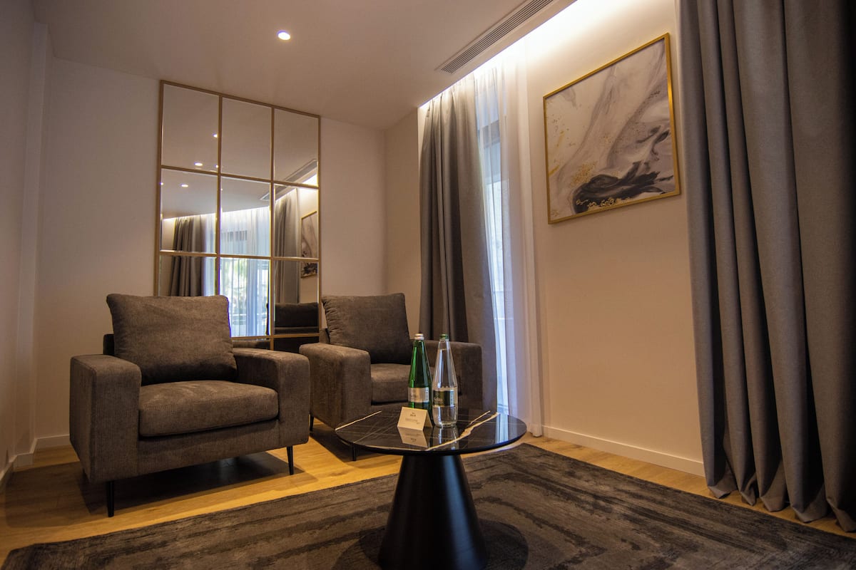 A modern hotel room with a seating area, a glass-top table, and a large mirror.