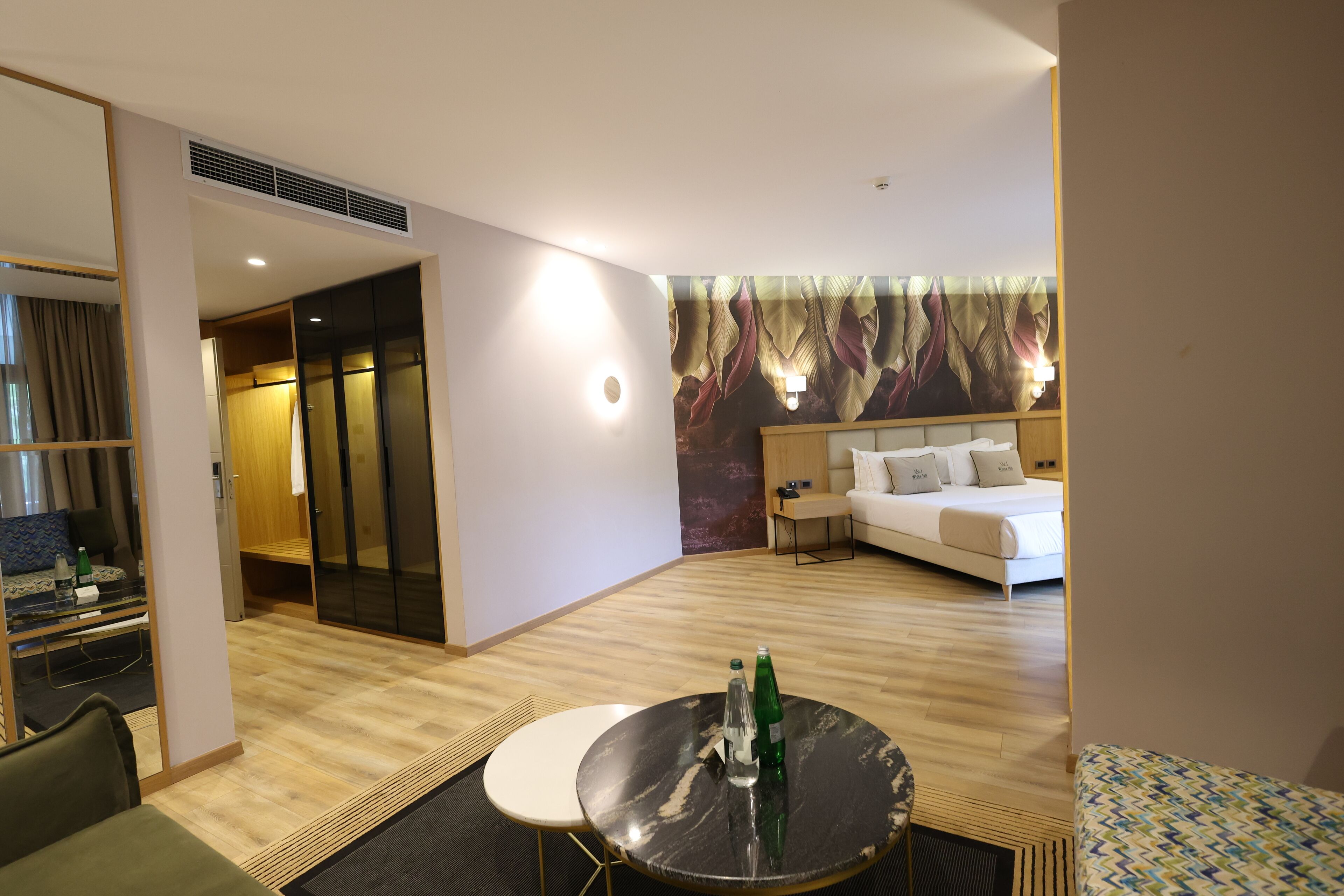 Habitación de hotel con cama, una mesa redonda con botellas, un sofá, un escritorio con una silla y un espejo grande.