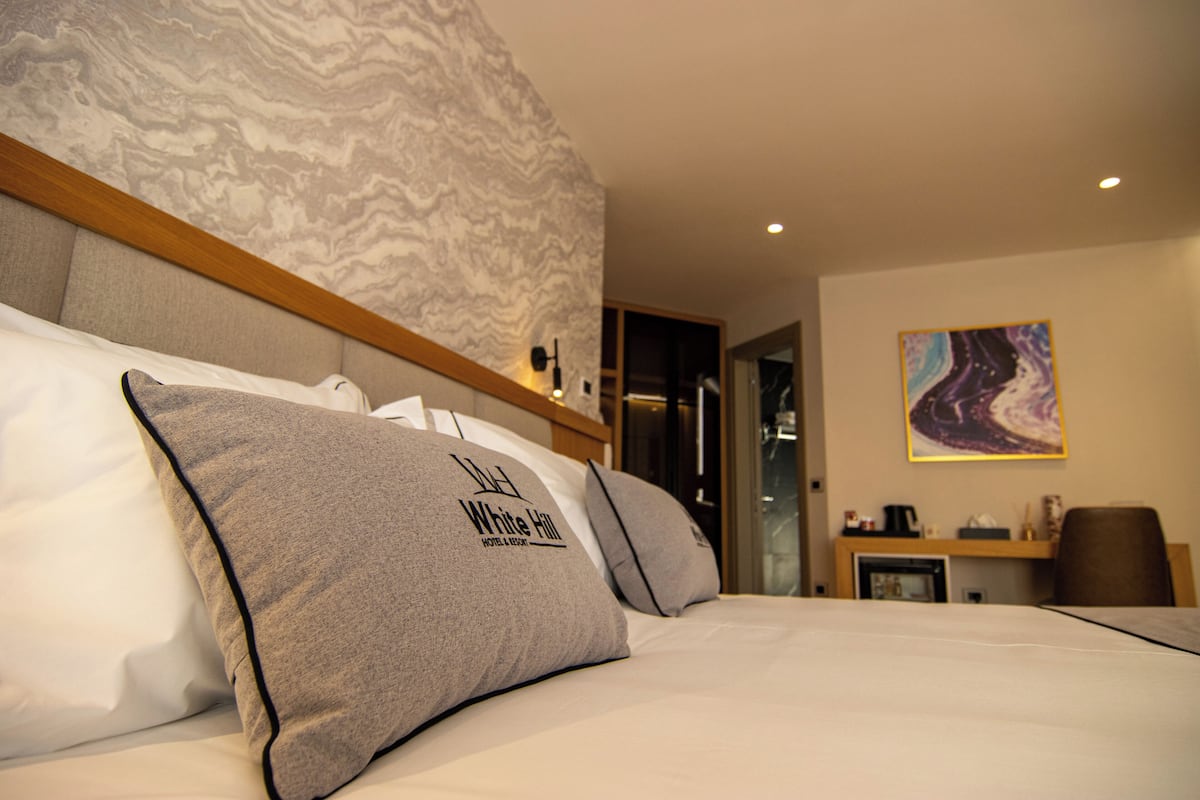 A hotel room with a bed, pillows, a headboard, a painting on the wall, and a desk with a chair.