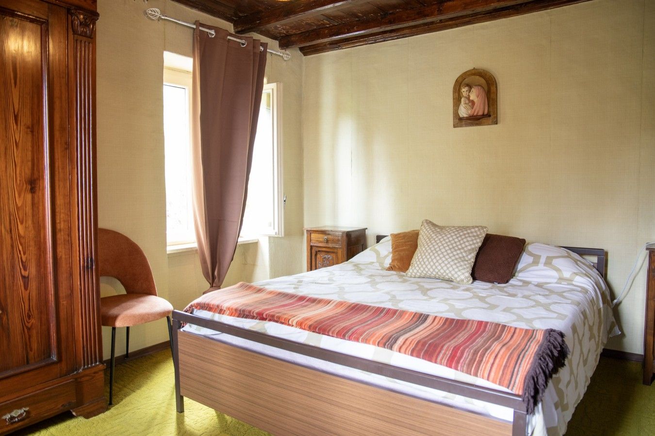 A bedroom with a wooden bed, a chair, a nightstand, and a religious painting on the wall.