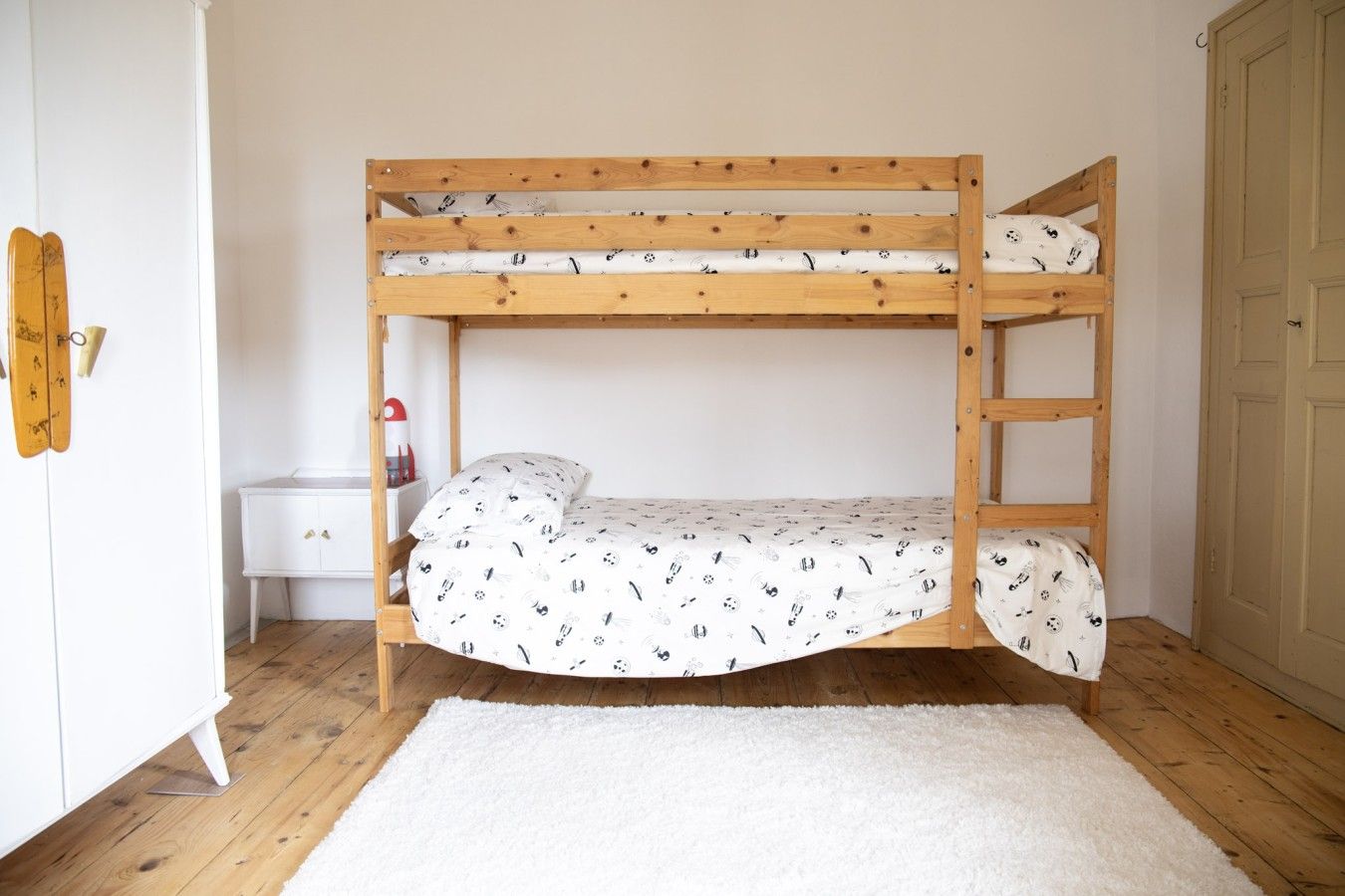 Ein Etagenbett mit weißer Matratze und gemusterten Betttextilien, ein weißer Nachttisch und ein weißer Kleiderschrank.