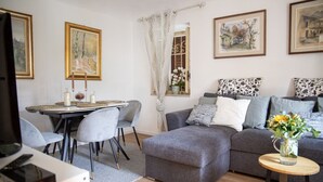 Un salon moderne avec un canapé gris, une table à manger avec des chaises et un téléviseur. Les murs sont ornés de tableaux encadrés, et un vase rempli de fleurs est posé sur une table d’appoint.