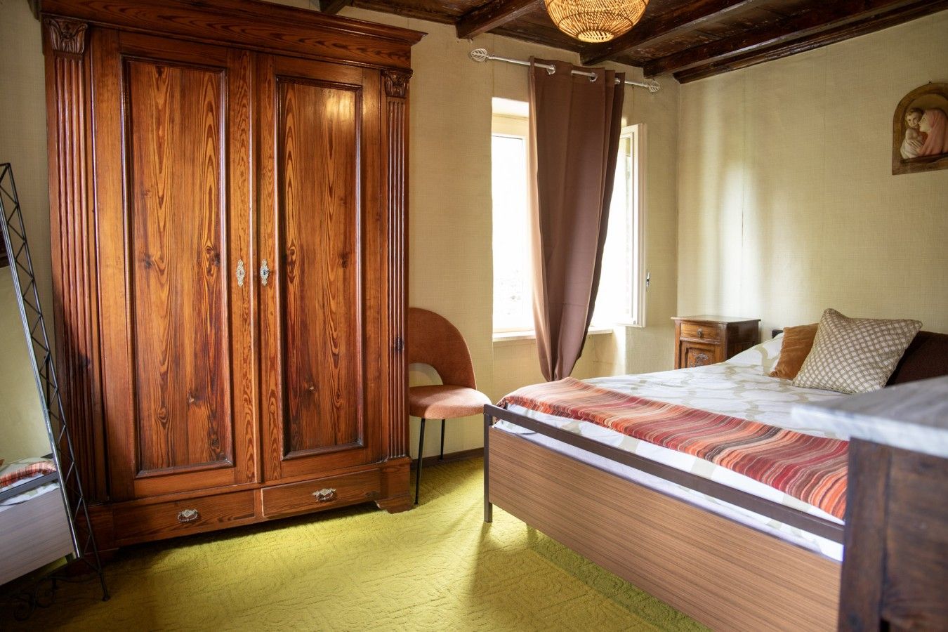 A bedroom with a wooden wardrobe, a bed with patterned bedding, a chair, and a window with curtains.