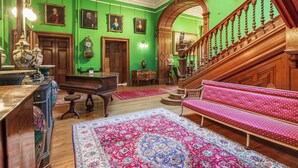 A grand room with green walls, a grand piano, ornate furniture, and a staircase with pink upholstery.