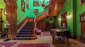 A grand foyer with a wooden staircase, a piano, and a green wall adorned with framed pictures.