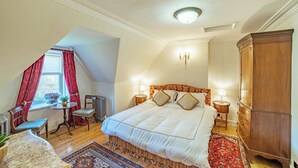 A bedroom with a large bed, a wooden wardrobe, a small table with a chair, and a window with red curtains.