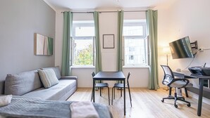 A modern living room with a sofa, a dining table with chairs, a desk with a computer and chair, and large windows with curtains. - Spacious 1BR Apt Near Danube (Vienna)