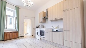 A modern kitchen with light wood cabinets, a built-in oven, a washing machine, and a window with green curtains. - Spacious 1BR Apt Near Danube (Vienna)