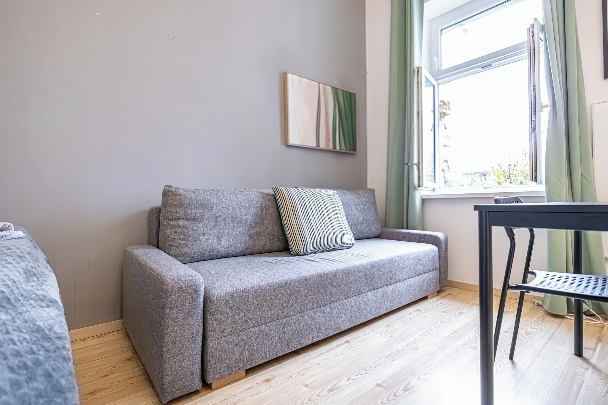 Un canapé gris avec un coussin à rayures, une petite table avec une chaise et une fenêtre avec des rideaux.