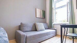 A grey sofa with a striped pillow, a small table with a chair, and a window with curtains. - Spacious 1BR Apt Near Danube (Vienna)