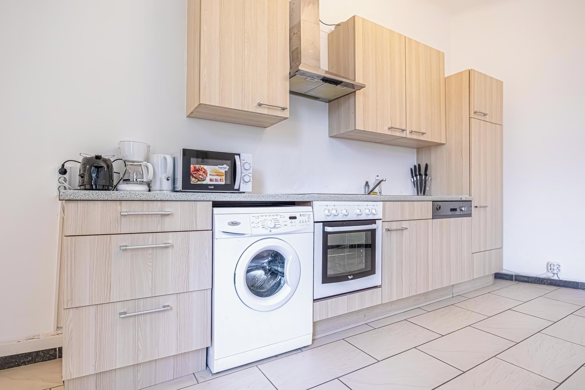 A modern kitchen with wooden cabinets, a washing machine, a microwave, and a coffee maker.