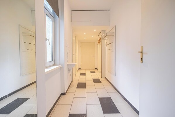 A narrow hallway with white walls, a checkered floor, and a window on the left.