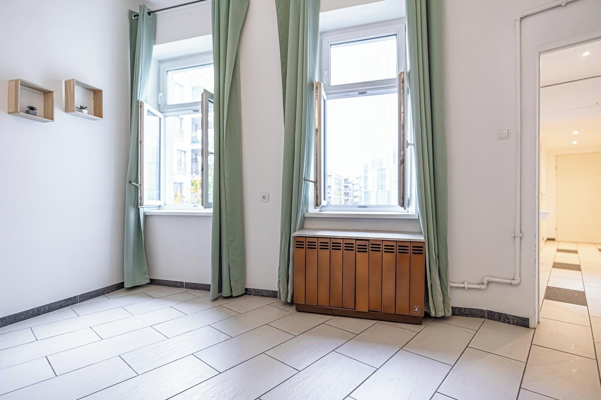 A room with two windows, a radiator, and a view of buildings outside.