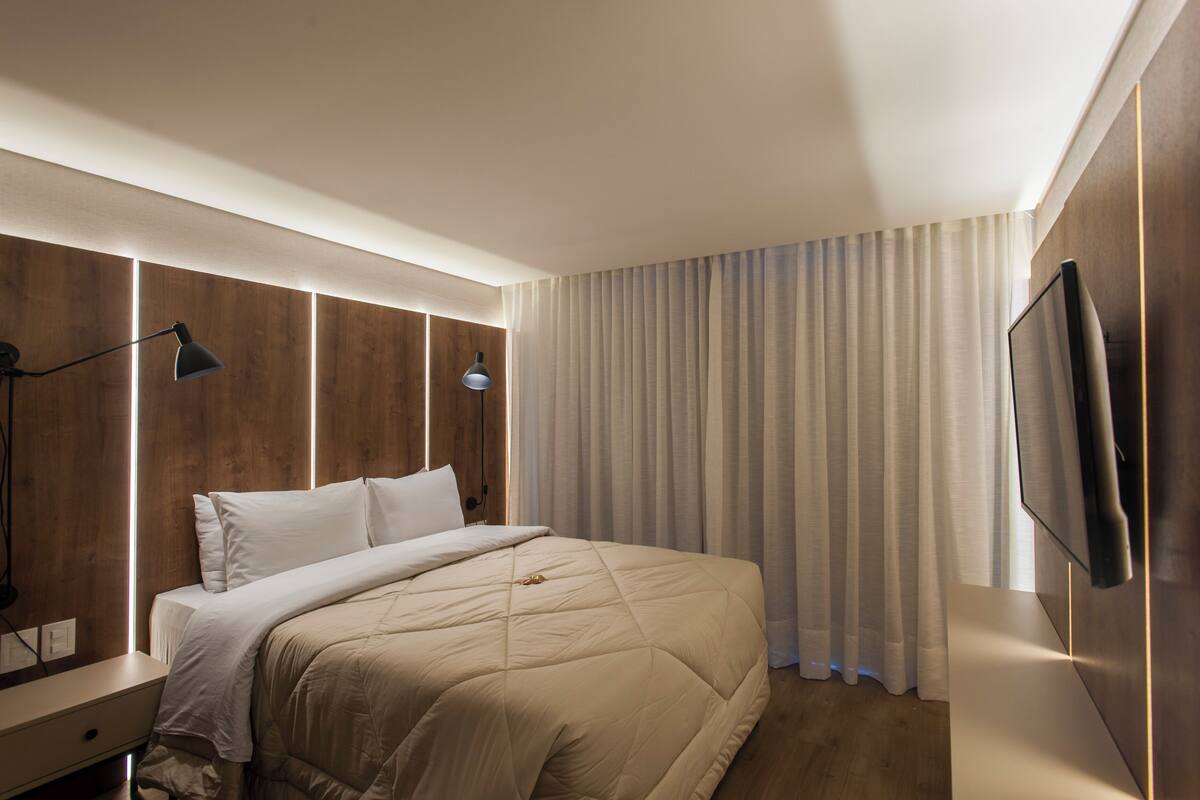 A modern hotel room with a large bed, wooden headboard, bedside lamps, a flat-screen TV, and a large window with curtains.
