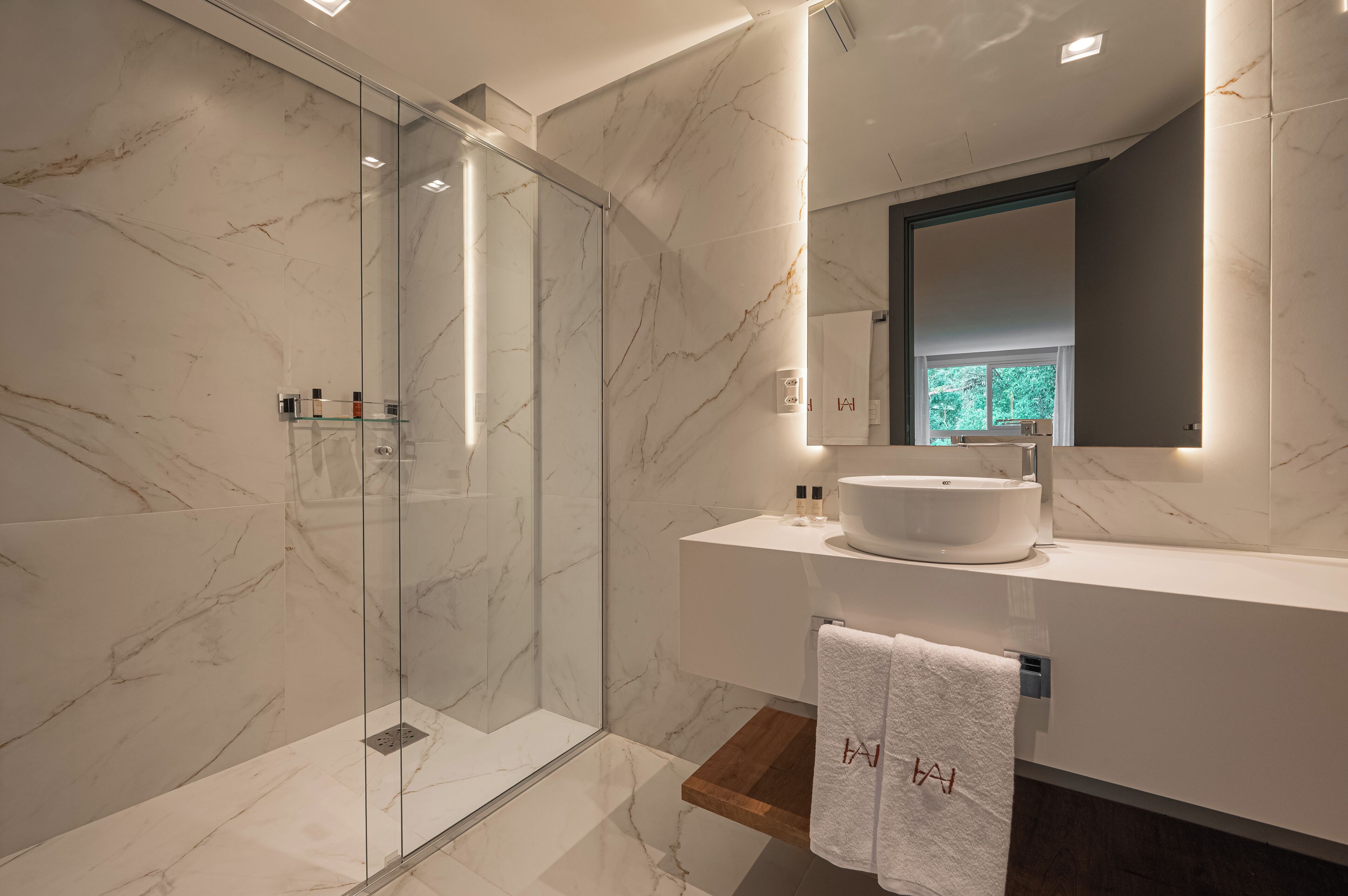 Un baño moderno con ducha envuelta en vidrio, pared con mosaicos de mármol, lavamanos blanco y estante de madera.