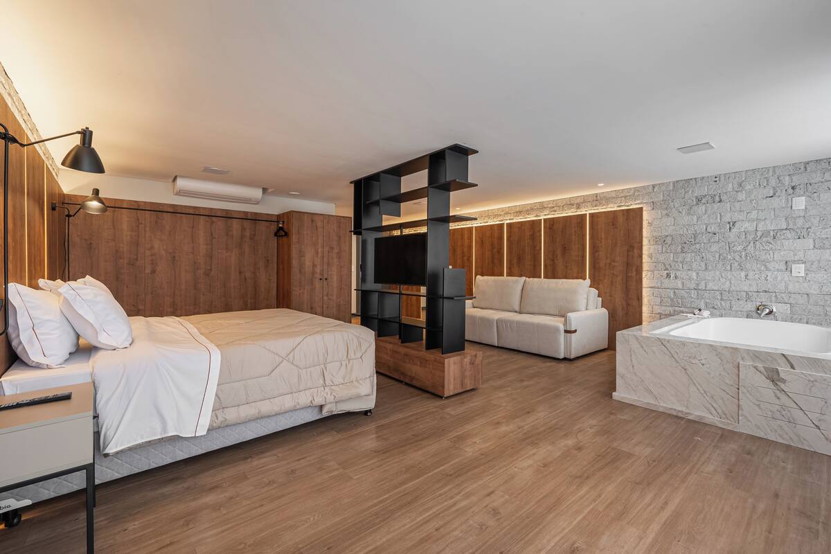 A modern bedroom with a large bed, a sofa, a built-in TV, a bathtub, and a stone wall.