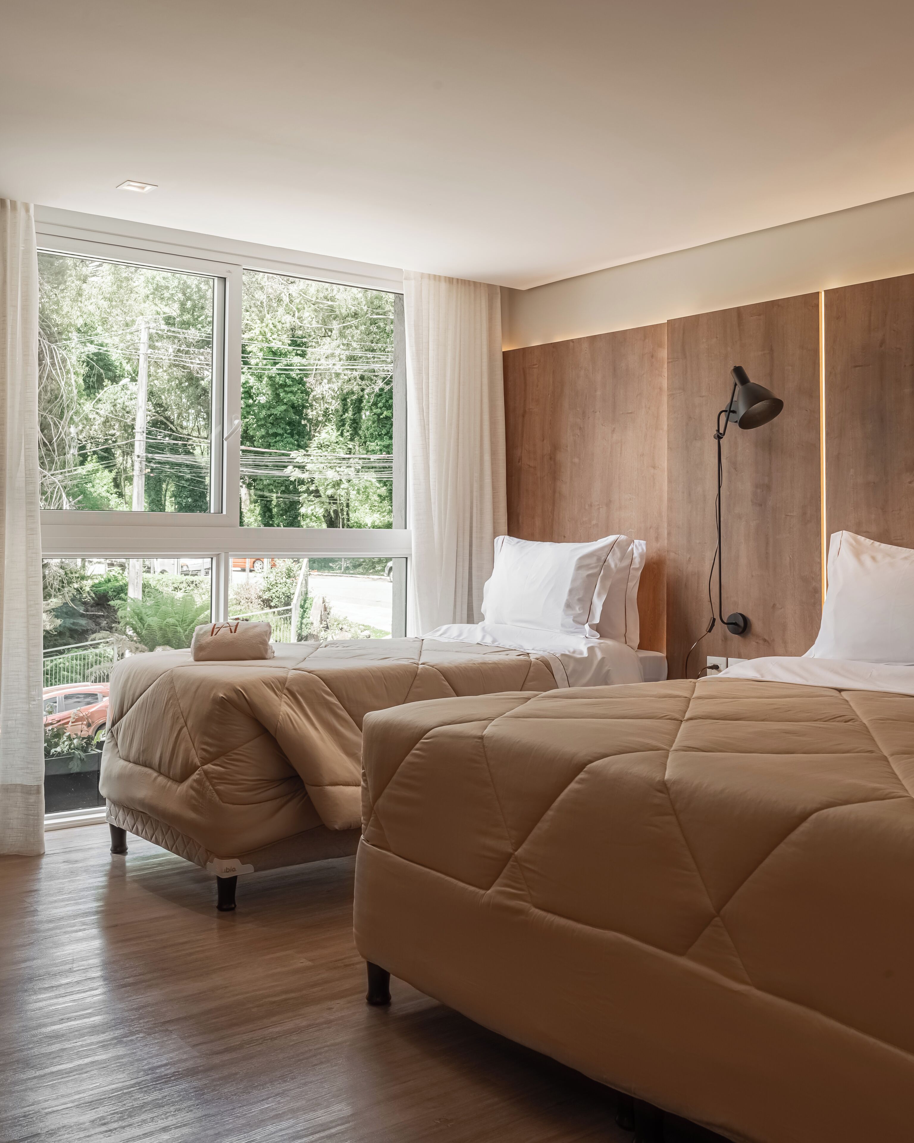 A modern hotel room with two beds, large windows, a wooden accent wall, and a nightstand with a lamp.