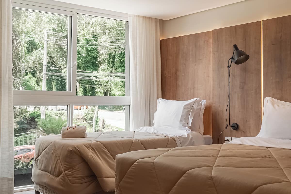 A modern hotel room with two beds, large windows, a wooden accent wall, and a nightstand with a lamp.