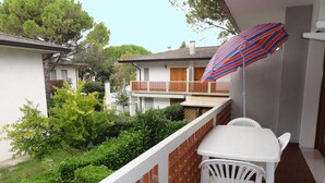 A balcony with a white table and chairs, a striped umbrella, and a view of neighboring buildings and greenery. - Apartment in a villa with balcony (Bibione)