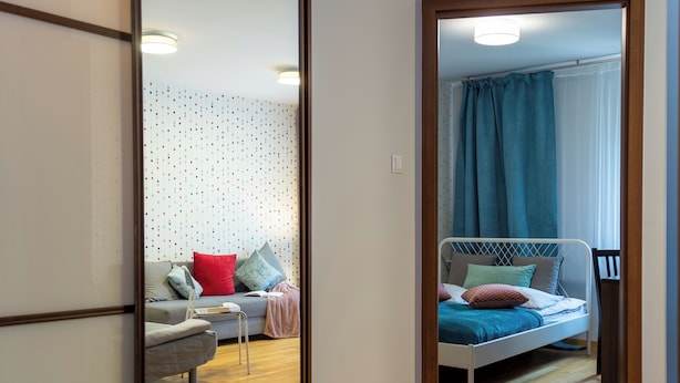 A modern bedroom with a wooden floor, a bed with blue bedding, a grey sofa with red and blue cushions, and a small table with a book. - AS HOME Nożownicza (Wrocław)