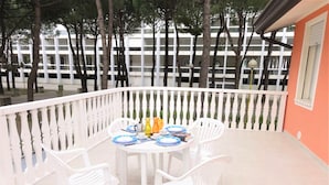 A balcony with a white railing, a round table with white chairs, and a view of a building with multiple windows. - Apartment in villa with fenced garden (Bibione)
