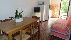 A compact room with a wooden dining table, chairs, a small kitchenette with a microwave and refrigerator, a TV, and a red metal bed frame. - Flat in a villa with private garden (Bibione)