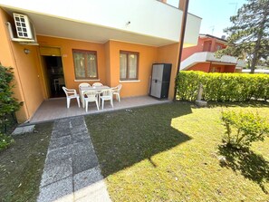A residential building with a patio area featuring a table and chairs, a door, and a small garden with a hedge.
