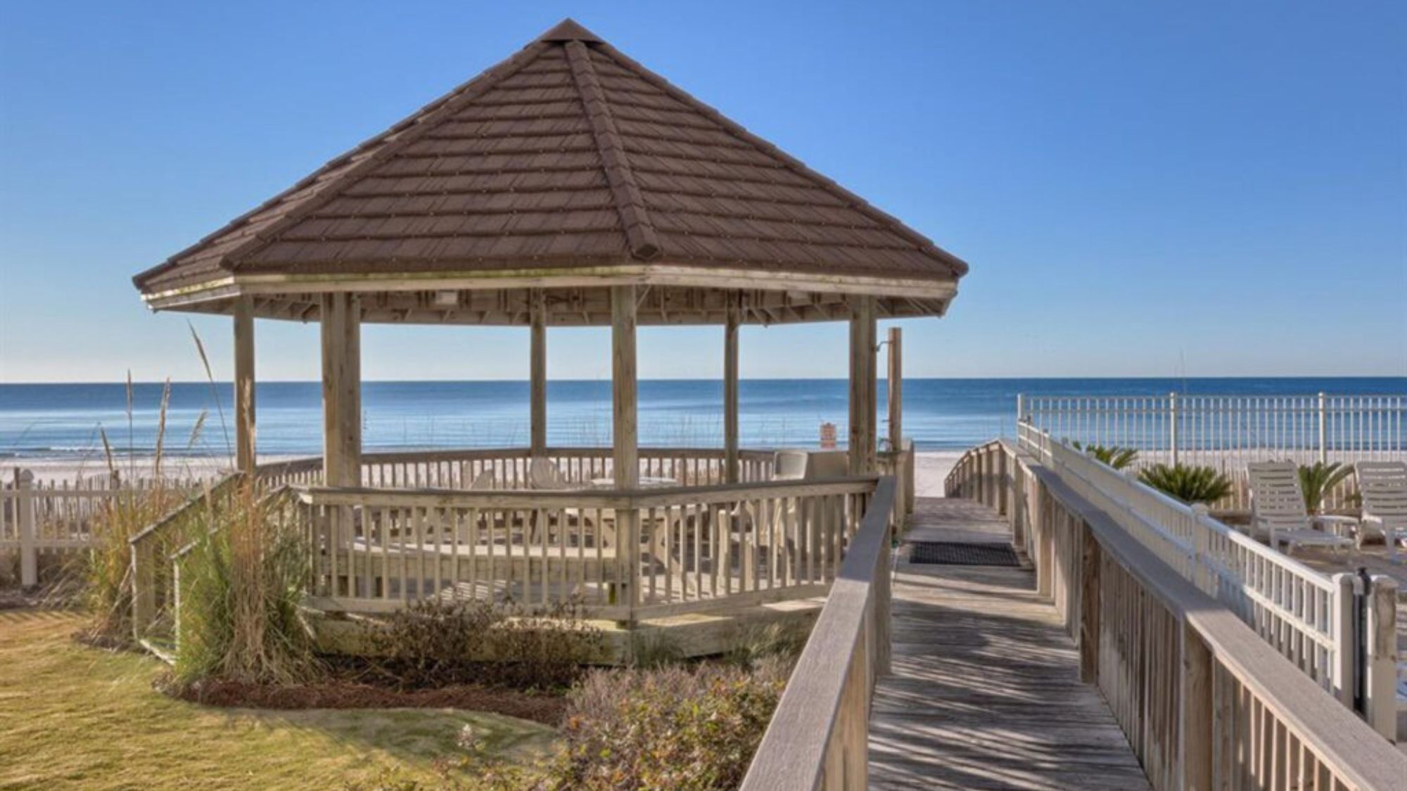 A wooden gazebo with a roof, surrounded by a wooden deck and railing, overlooking a beach.