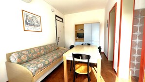 A compact living space with a sofa, a dining table, and chairs. There is a kitchenette area with cabinets and a television.