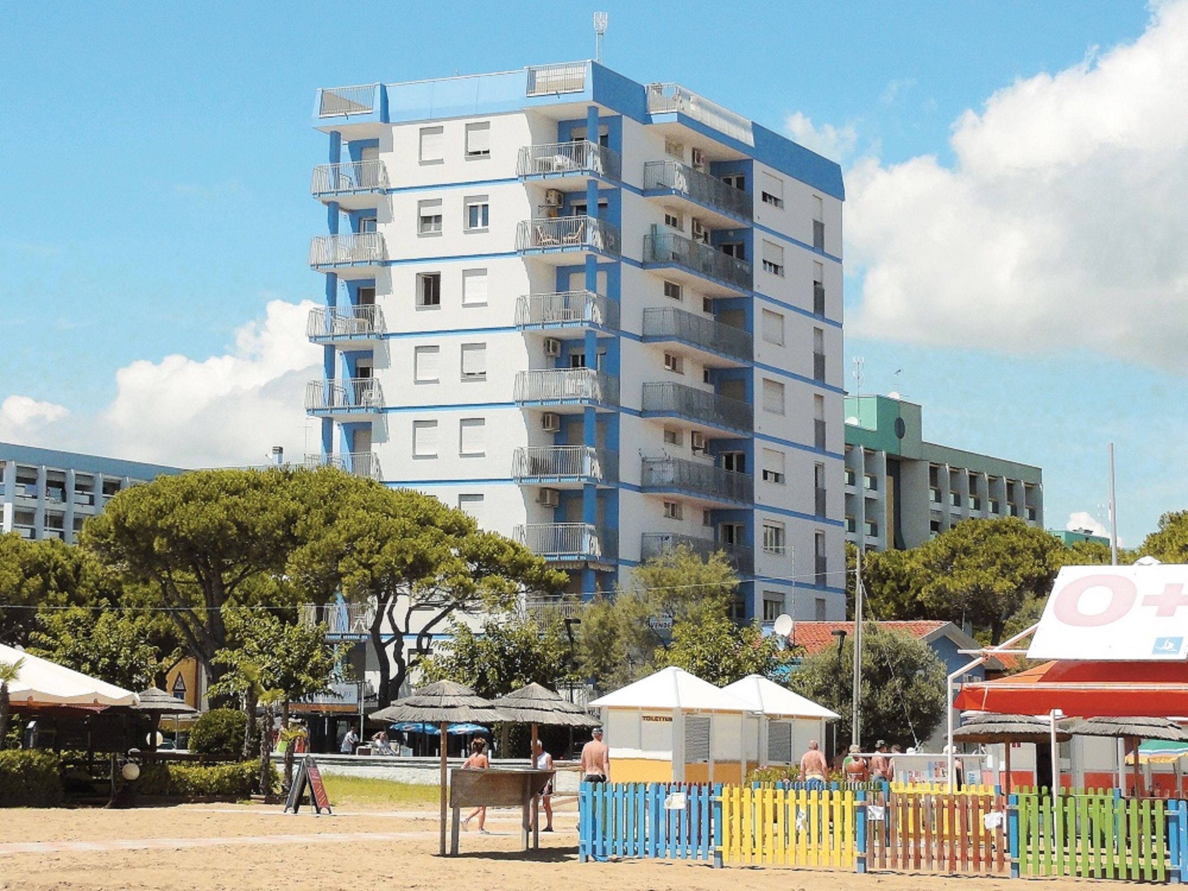 A multi-story building with balconies, a sandy ground, and some trees.