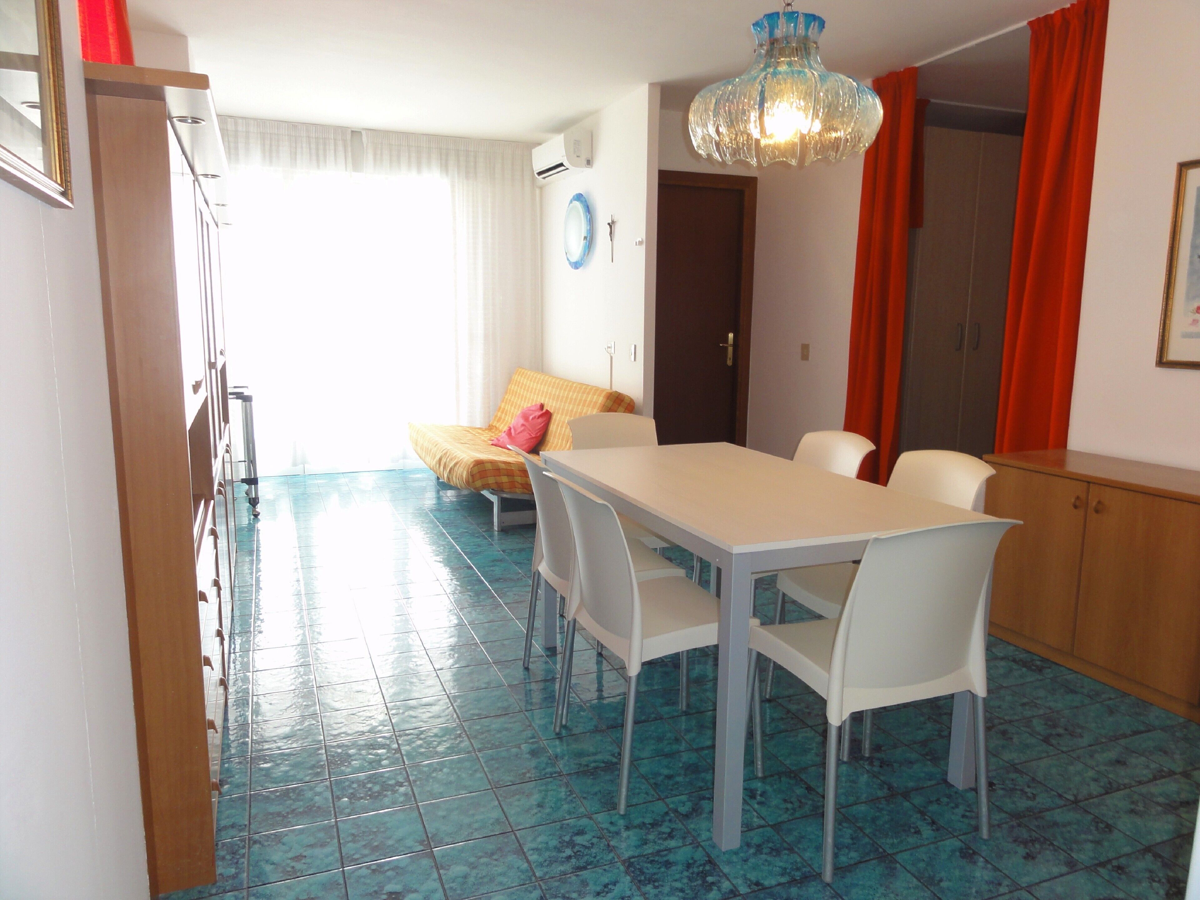 Un comedor con una mesa y sillas blancas, piso de baldosas azules, cama con colcha amarilla y cortina roja.