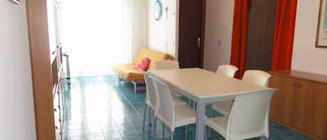 Un comedor con una mesa y sillas blancas, piso de baldosas azules, cama con colcha amarilla y cortina roja.