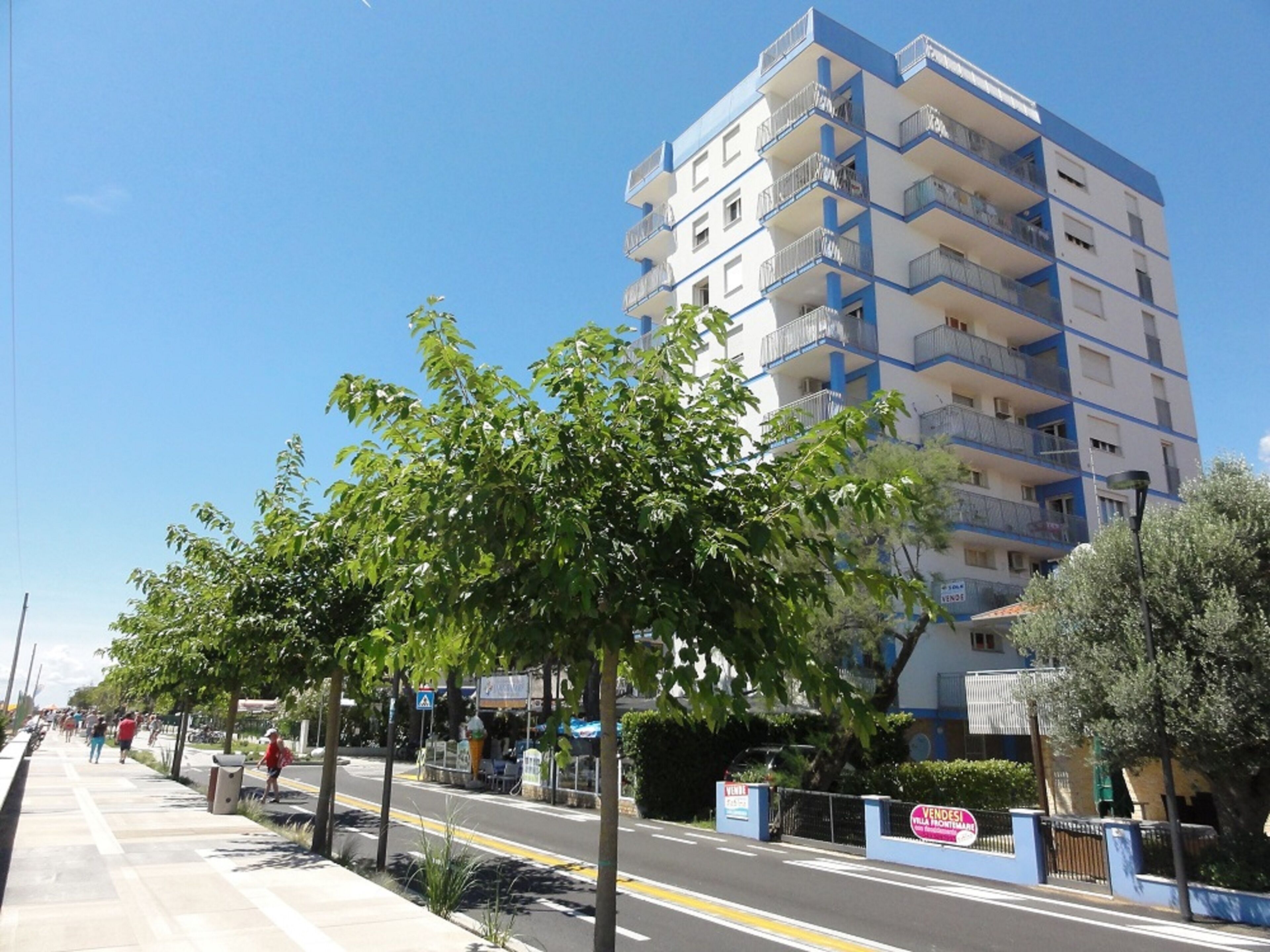 A multi-story building with balconies, a paved sidewalk, and trees lining the street.