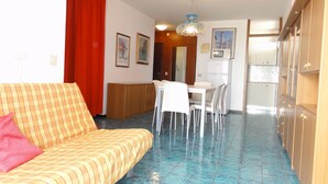 Ein Wohnzimmer mit einer Couch, einem Esstisch mit Stühlen, einer Küchenzeile mit Schränken und einem roten Vorhang.