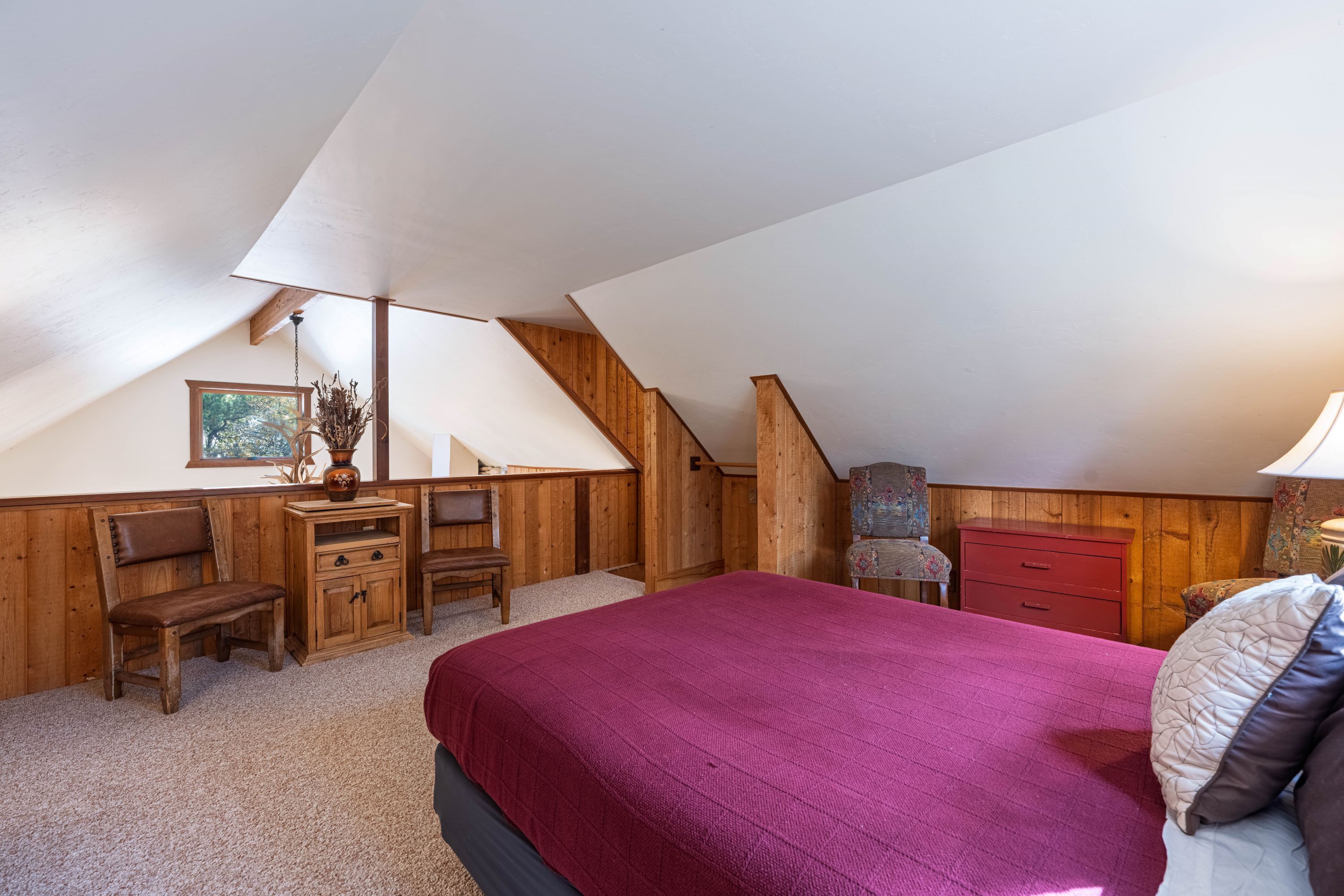 A bedroom with a sloped ceiling, wooden walls, a bed with a red blanket, a desk with a chair, and a dresser.