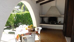 An outdoor patio with a table set for dining, surrounded by white chairs, a barbecue grill, and a basket of sunflowers.