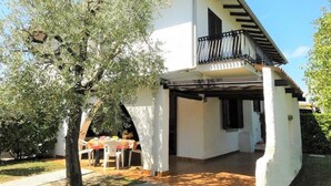 A white building with a balcony, a tree in front, and a table with chairs outside.
