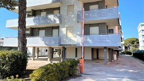 A multi-story building with balconies, a red pole, and a paved area with bushes. - Flat in seafront condominium with parking (Bibione)