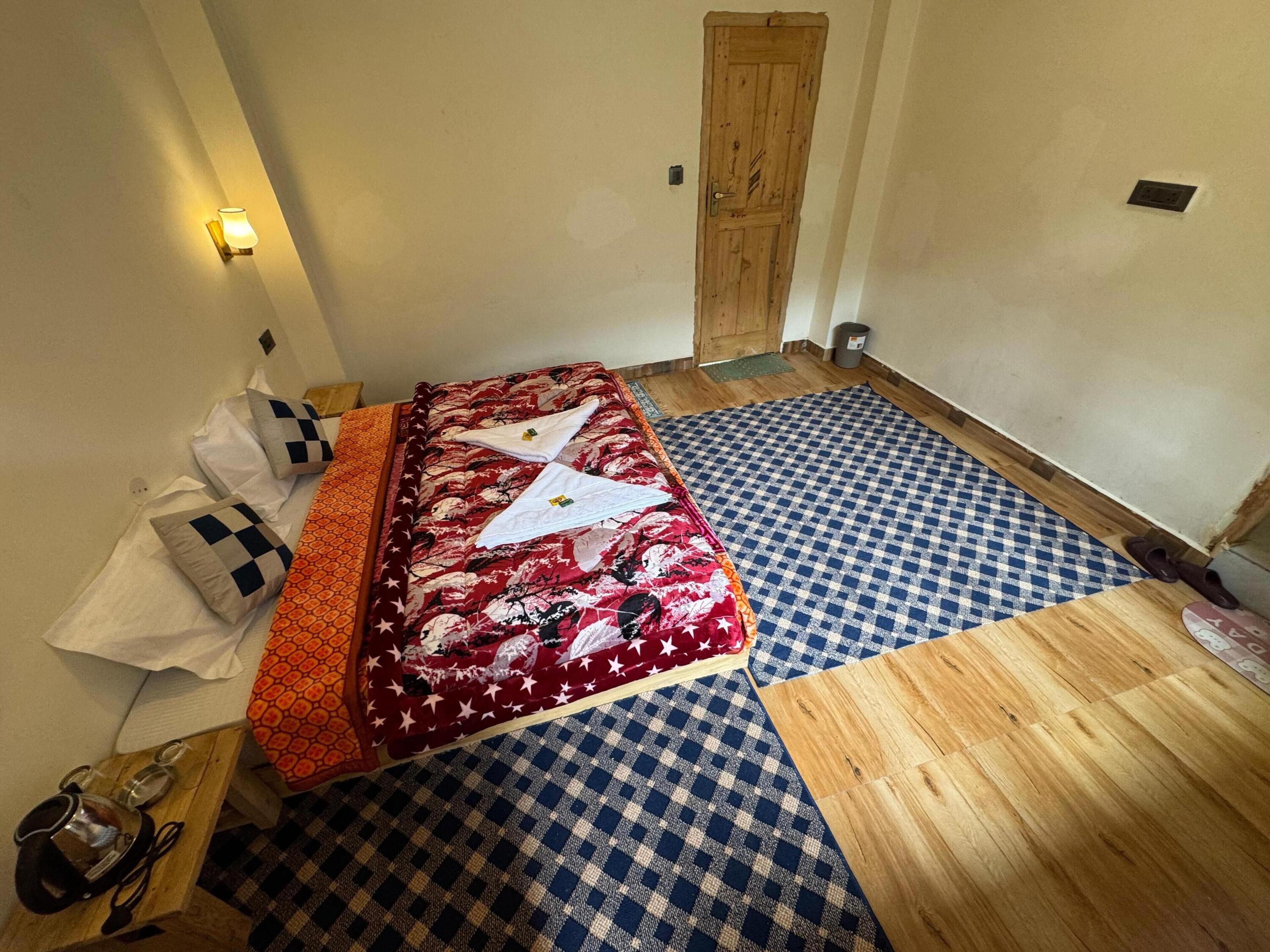 A room with a bed, a wooden floor, a checkered rug, and a wall-mounted light.