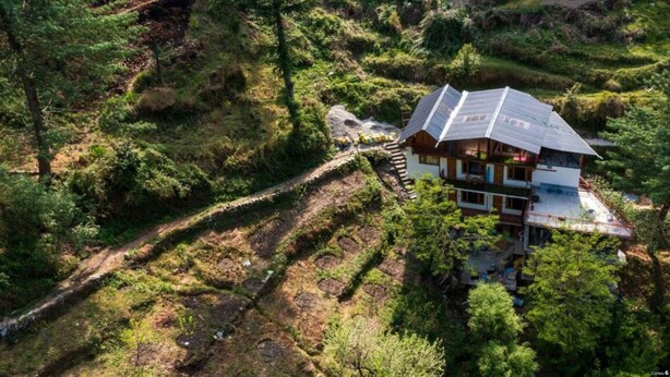 Une maison en bois à deux étages, dotée d’un toit en pente, entourée de champs en terrasses et d’une forêt dense.