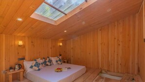 Une chambre dans une cabane en bois, dotée d’une lucarne, d’un lit agrémenté d’oreillers à motifs, d’une table de chevet avec une lampe et d’une petite table sur laquelle se trouvent une bouilloire et un panier de fleurs.