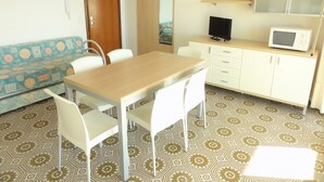 Ein Zimmer mit einem Holztisch, weißen Stühlen, einem Sofa mit gemustertem Bezug, einem Schrank mit Mikrowelle und einem Spiegel an der Wand.