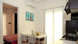 A compact hotel room with a kitchenette, a dining area with a table and chairs, a bed with a colorful bedspread, and a painting on the wall.