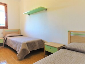 Ein schlichtes Zimmer mit zwei Betten, einem grünen Regal und einem Fenster.