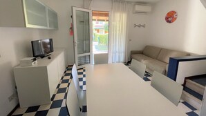 Een moderne, goed verlichte kamer met een zwart-wit betegelde vloer, een witte tafel, een bank, een televisie en een schuifdeur naar een balkon.