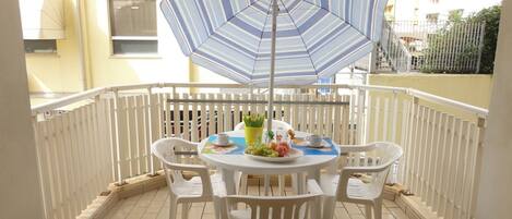 A balcony with a round table, white plastic chairs, a striped umbrella, and a small potted plant.