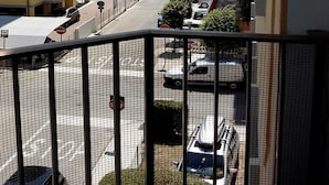 Une vue depuis un balcon donnant sur une rue où sont garées des voitures, un parking et des bâtiments en arrière-plan.