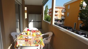 Een balkon met een tafel gedekt voor twee personen, witte stoelen en uitzicht op de straat en gebouwen.