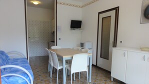 A small, compact room with a bed, a dining table with chairs, a television mounted on the wall, and a cabinet.